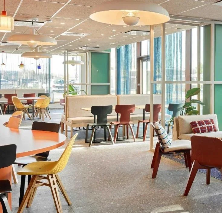 Intérieur lumineux d'un café ou d'un espace de coworking avec plusieurs tables et chaises colorées, grandes fenêtres laissant entrer la lumière naturelle, et un espace lounge avec canapé et fauteuils.