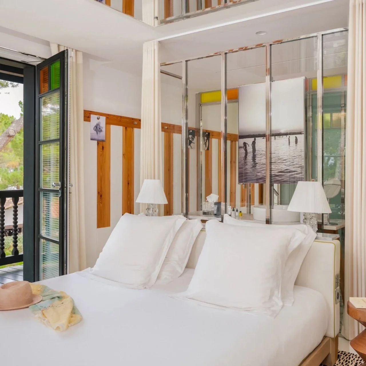 Chambre d'hôtel avec un lit blanc, lampes de nuit, et une fenêtre ou porte-fenêtre ouverte donnant sur un balcon avec vue sur des arbres et un paysage extérieur, décorée avec des photos ou œuvres d'art.