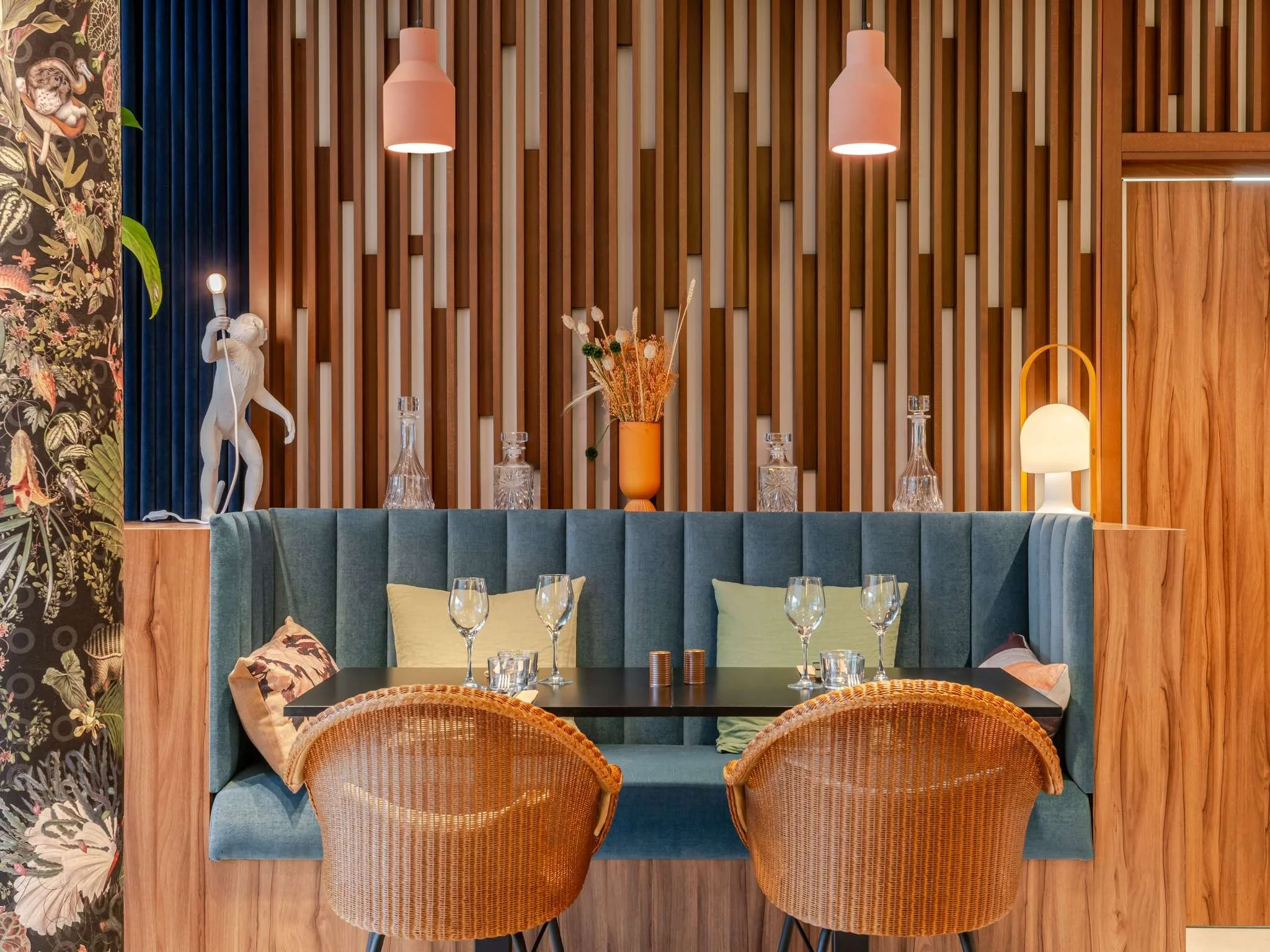 Intérieur d'un restaurant avec un mur en bois, des lampes suspendues, un vase avec des fleurs, une banquette en velours bleu, une table dressée avec des verres et des coussins sur les sièges.