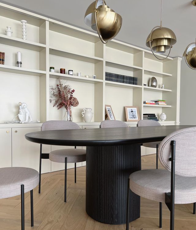 Salle à manger avec une table ronde noire et des chaises en tissu gris clair, bibliothèque blanche en arrière-plan avec décorations et livres, suspendus en plafonnier en métal brillant doré.