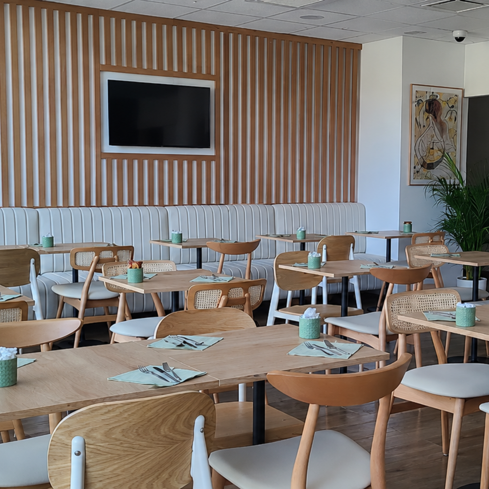 Salle à manger d'un restaurant avec des tables en bois, des chaises en bois, un mur en bois avec une télévision, une peinture de femme africaine, et une grande plante verte.