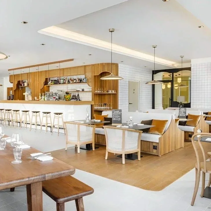 Intérieur d'un restaurant moderne avec bar en bois, tabourets de bar, tables en bois, chaises blanches, et murs en carreaux blancs, ambiance lumineuse et accueillante.