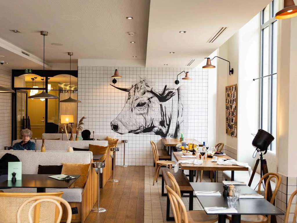 Intérieur d'un restaurant avec des tables, des chaises en bois, un mur avec une grande fresque d'un taureau, et une grande fenêtre laissant passer la lumière.