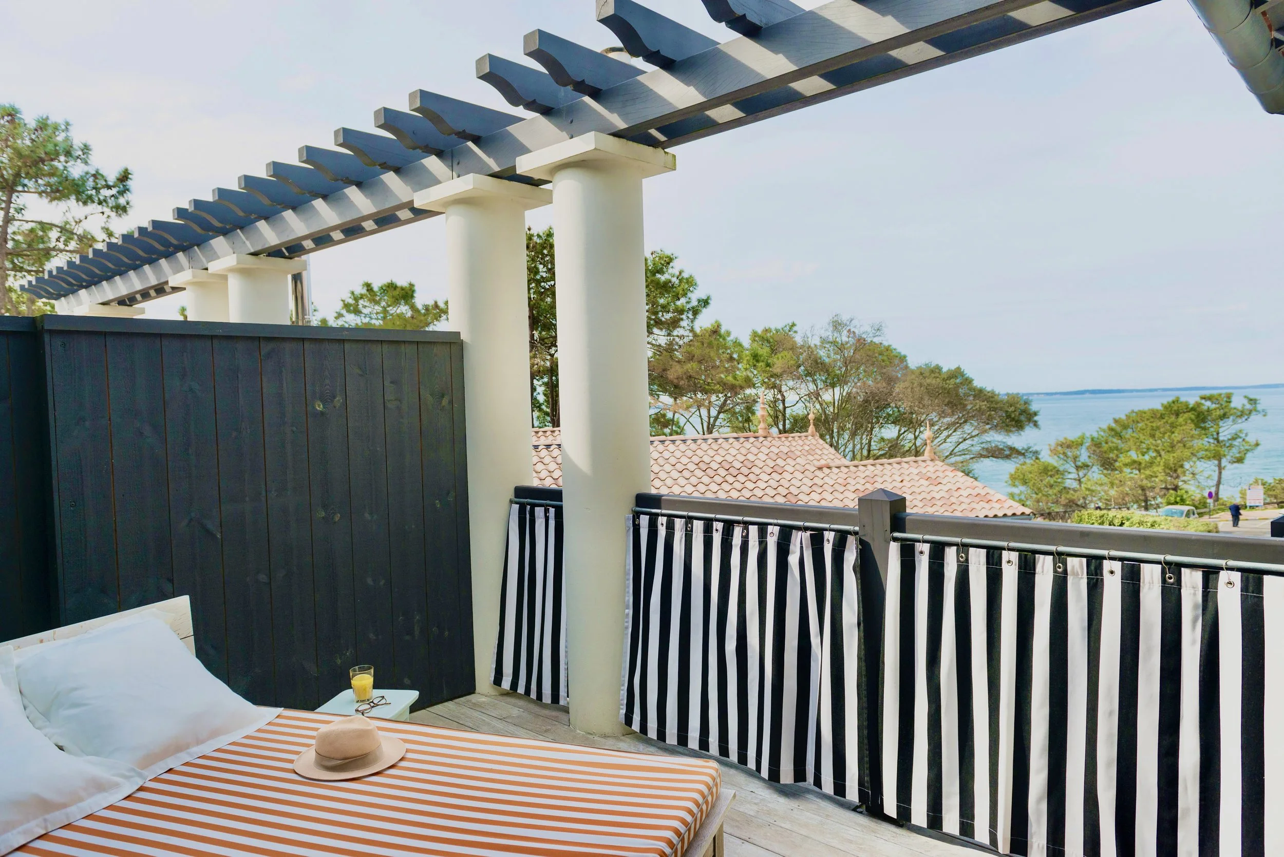Une terrasse d'hôtel ou de résidence avec vue sur la mer, comprenant un lit avec une serviette de plage, un chapeau, une boisson dans un verre, et un matelas ou canapé noir avec rayures blanches contre une clôture en bois peint en noir, entourée de p