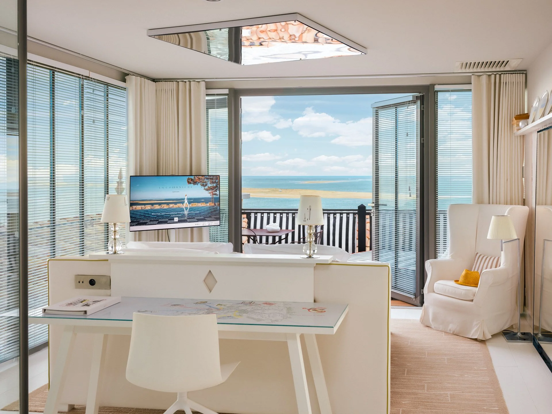 Salon avec vue sur la mer, grands rideaux blancs, fauteuils confortables, télévision, table avec carte et livre.