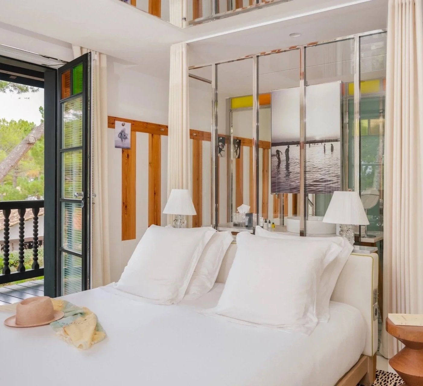 Une chambre d'hôtel lumineuse avec un lit blanc, lampes de chevet, une porte-fenêtre ouverte donnant sur un balcon avec vue sur des arbres, un tableau représentant des personnes dans l'eau, avec des rideaux blancs et un miroir intégré.