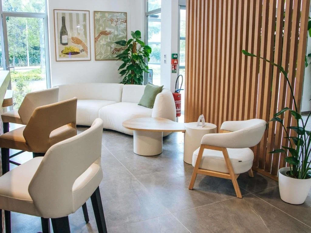 Salon de réception avec canapé blanc, fauteuils beige et blanc, table basse ronde en bois clair, décor avec plantes vertes et œuvres d'art au mur, séparateur en bois, grande fenêtre, sol en carreaux gris, ambiance moderne et lumineuse.