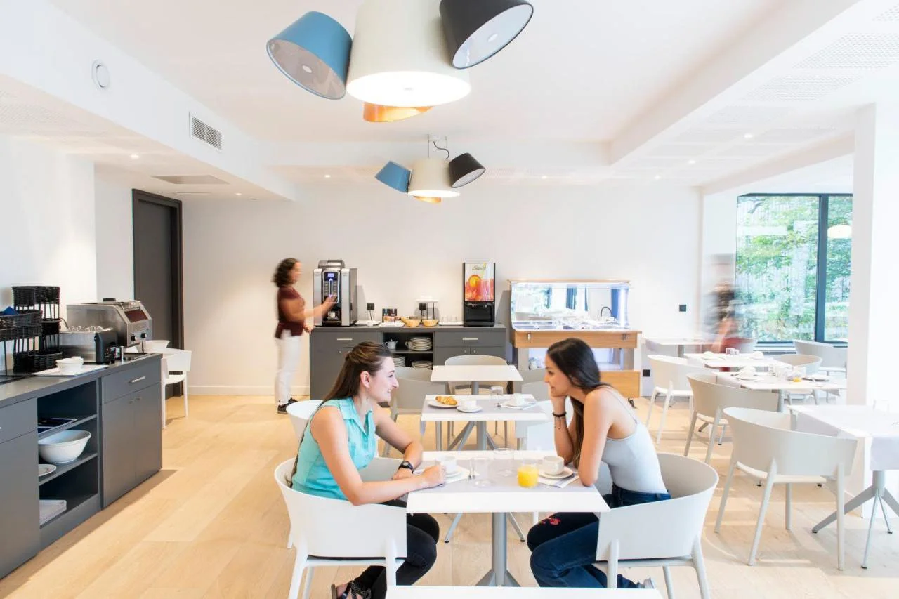 Deux femmes discutent à une table dans une salle de petit-déjeuner lumineuse. Une femme en cuir blanc et une autre en turquoise, un verre de jus d'orange devant elles. À l'arrière, un buffet avec machines à café et autres aliments, et une femme se se