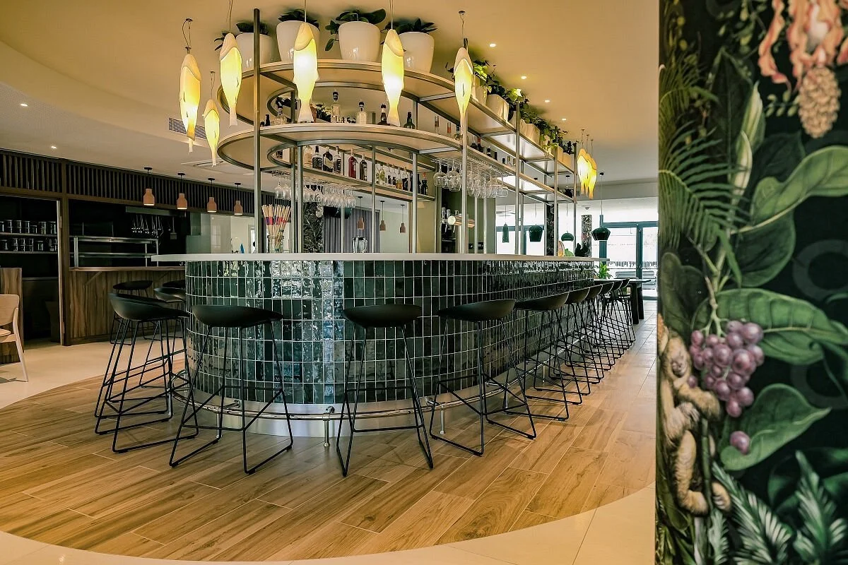 Un bar moderne avec un comptoir en mosaïque verte, des chaises hautes noires, des lampes design suspendues, et des étagères avec des plantes et des bouteilles en arrière-plan