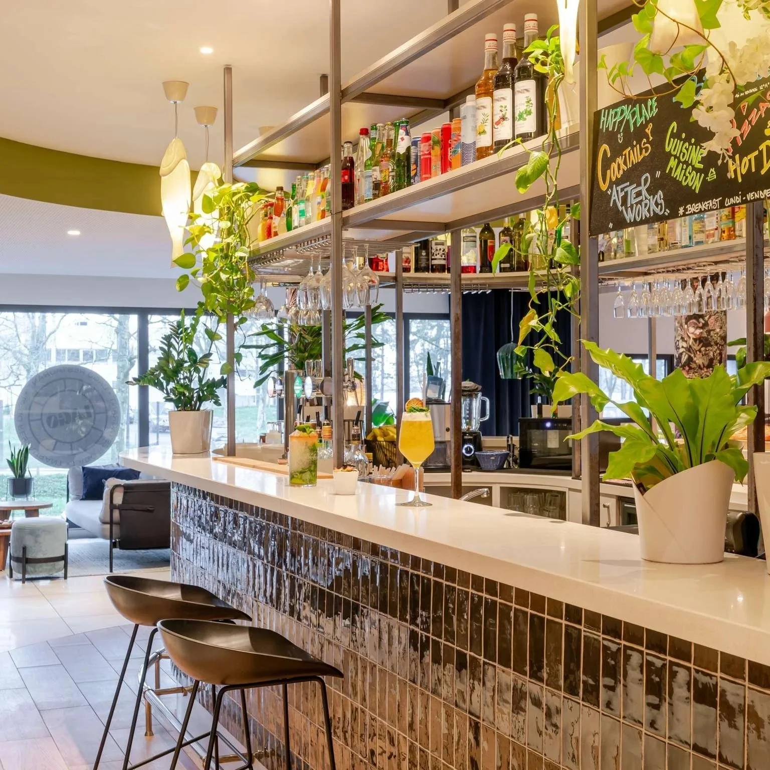 Bar moderne avec boissons colorées, plantes vertes, étagères de bouteilles d'alcool, comptoir en marbre, et tableau noir indiquant des cocktails et autres offres.