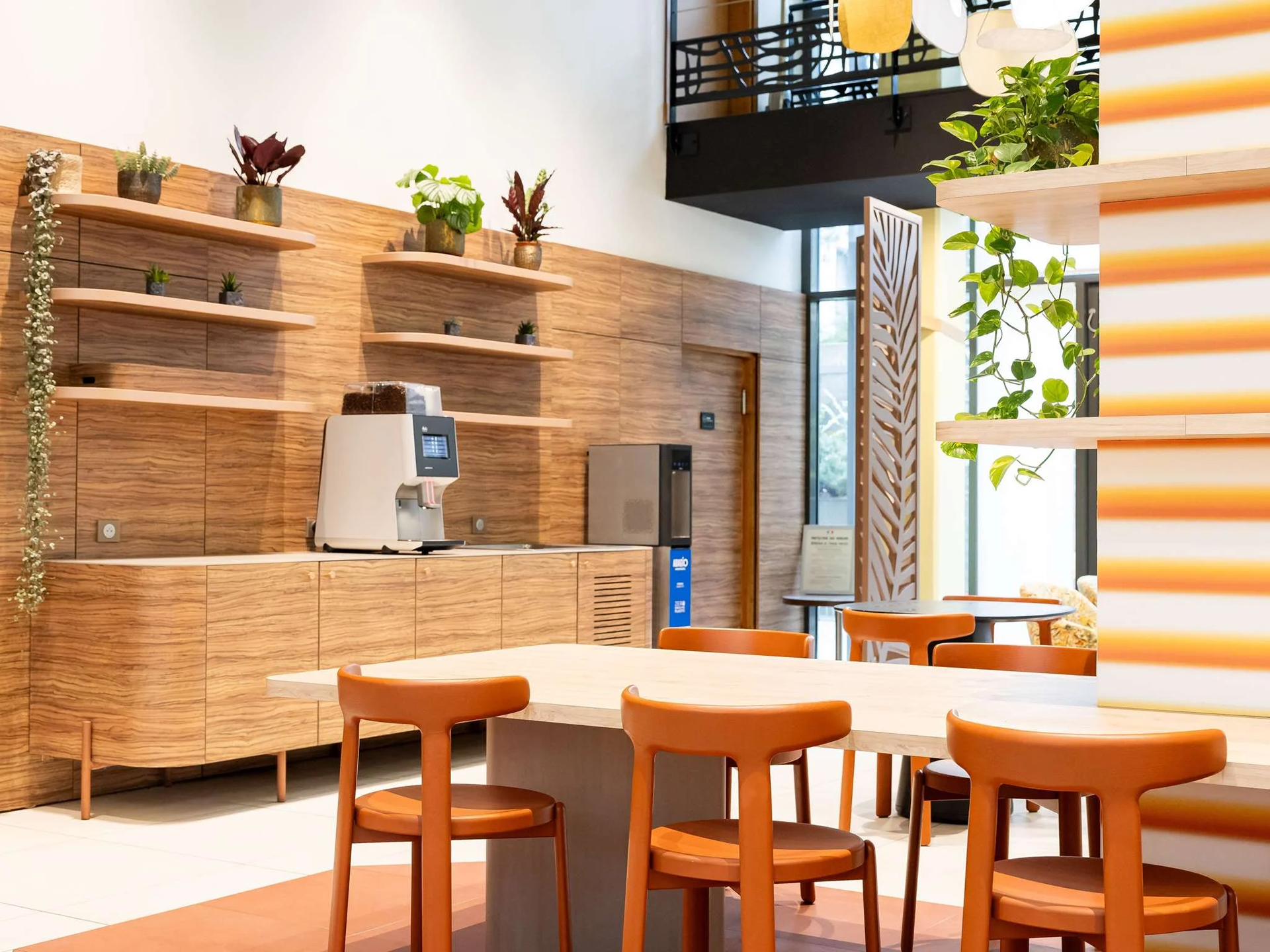 Intérieur d'une salle de restauration avec des chaises en bois orange, des tables blanches, des étagères en bois avec des plantes vertes et décoratives, et un mur en bois avec une grande fenêtre laissant entrer la lumière naturelle.
