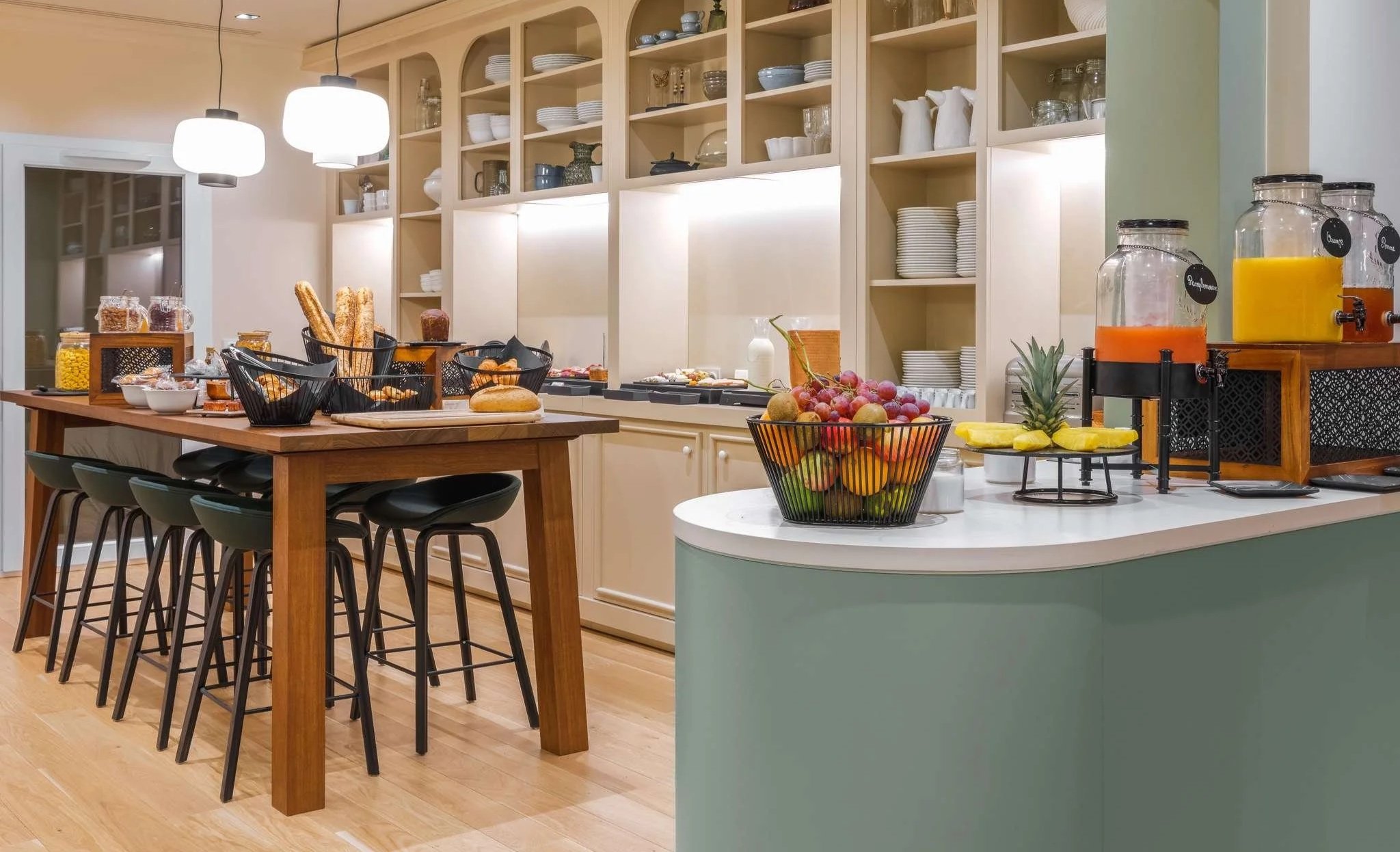 Cuisine moderne avec une table en bois et des chaises noires, étagères blanches avec vaisselle, fruits, et distributeurs de jus de fruits sur le comptoir.