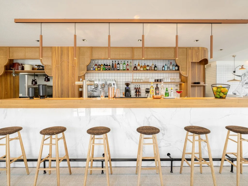 Un comptoir de bar en marbre blanc avec des tabourets en boismatériel clair, derrière un étagère en bois avec des bouteilles d'alcool, un miroir et des éléments de bar, dans un intérieur moderne et lumineux.