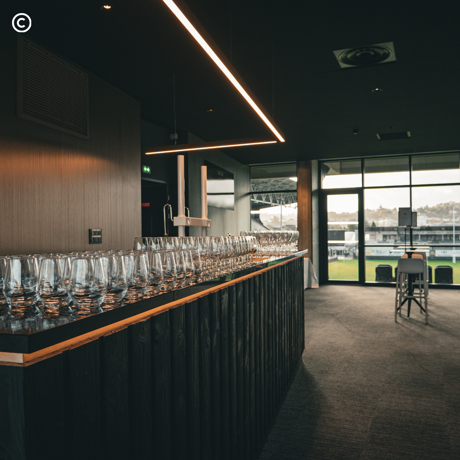Intérieur d'un bar ou restaurant moderne avec un comptoir en bois foncé, un mur en bois clair avec des tasses ou verres alignés, des plafonds noirs avec un éclairage linéaire chaud, de grandes fenêtres laissant passer la lumière naturelle et offrant 