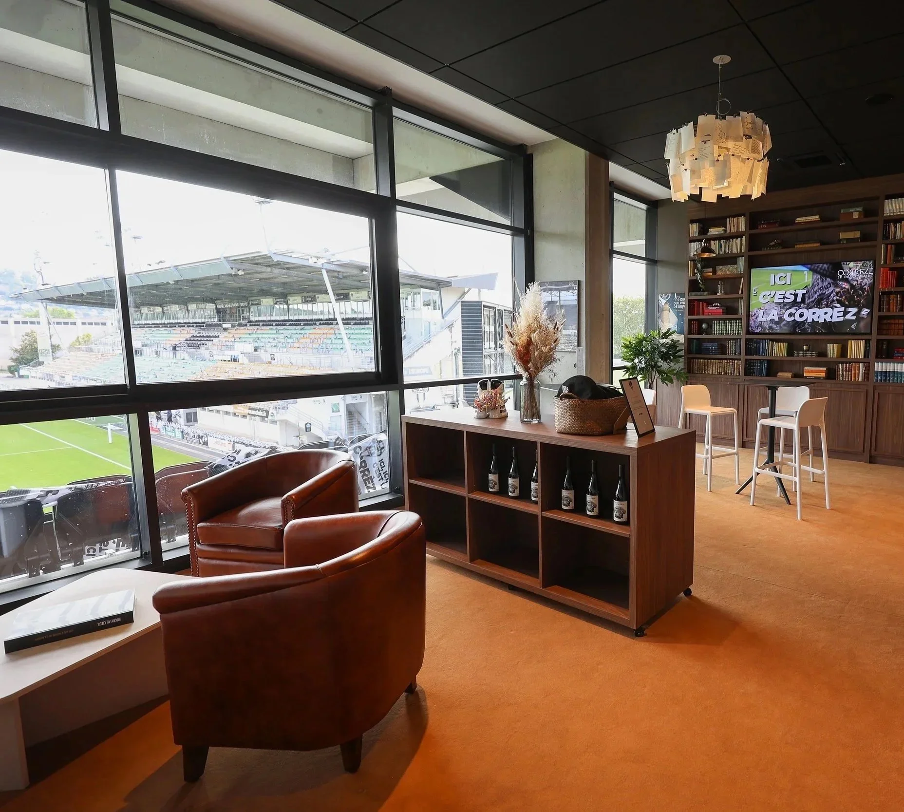 Intérieur d'une salle avec grande fenêtre donnant sur un stade. Mobilier comprenant deux fauteuils en cuir, une étagère avec des bouteilles, un panneau avec des livres, une télévision, et des décorations végétales.