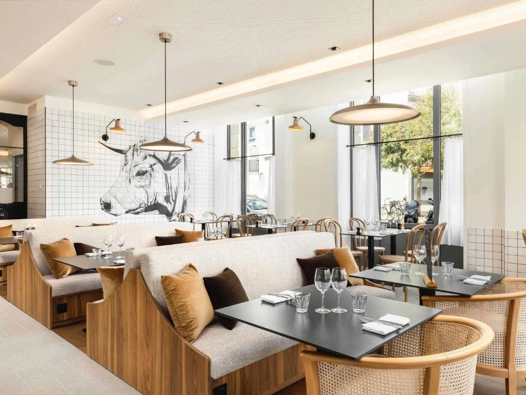 Intérieur d'un restaurant moderne avec tables, chaises en bois, coussins, grands fenêtres, mur avec dessin d'une tête de taureau en noir et blanc, éclairage suspendu.