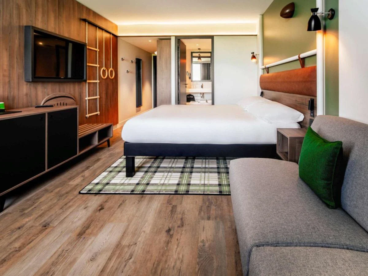 Chambre d'hôtel moderne avec lit double, télévision, fauteuil gris avec coussin vert, sol en bois, mur en bois clair, salle de bain visible au fond.
