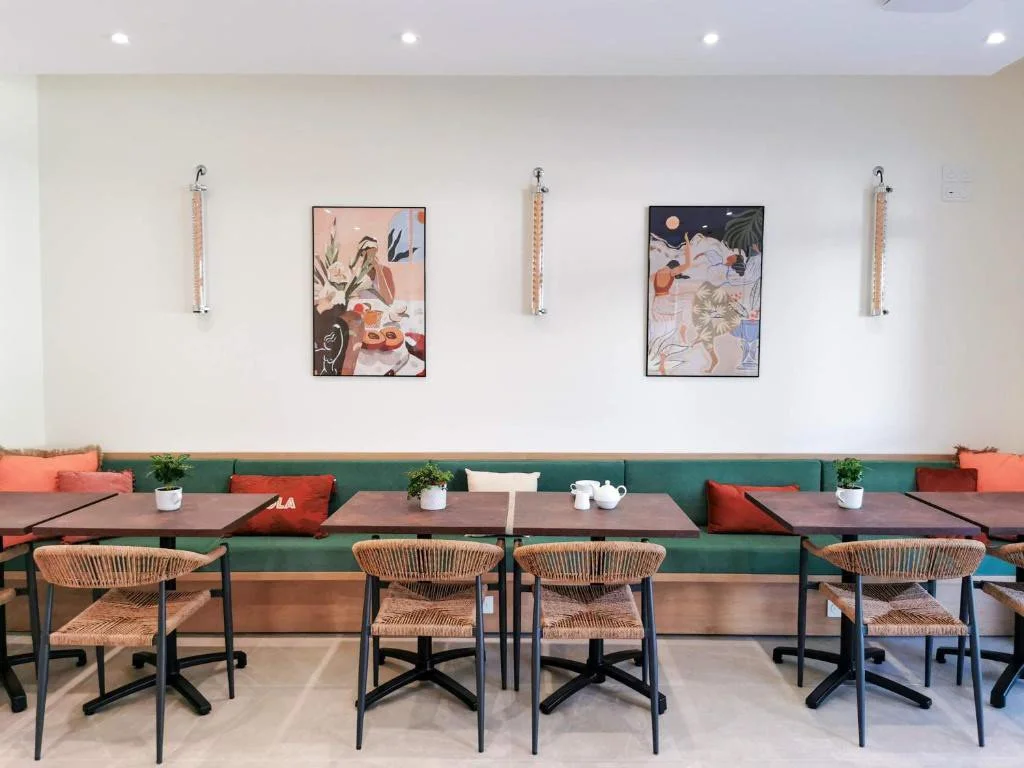 Intérieur d'un café avec une banquette verte, plusieurs tables en bois, des chaises en rotin, et des décorations murales abstraites.