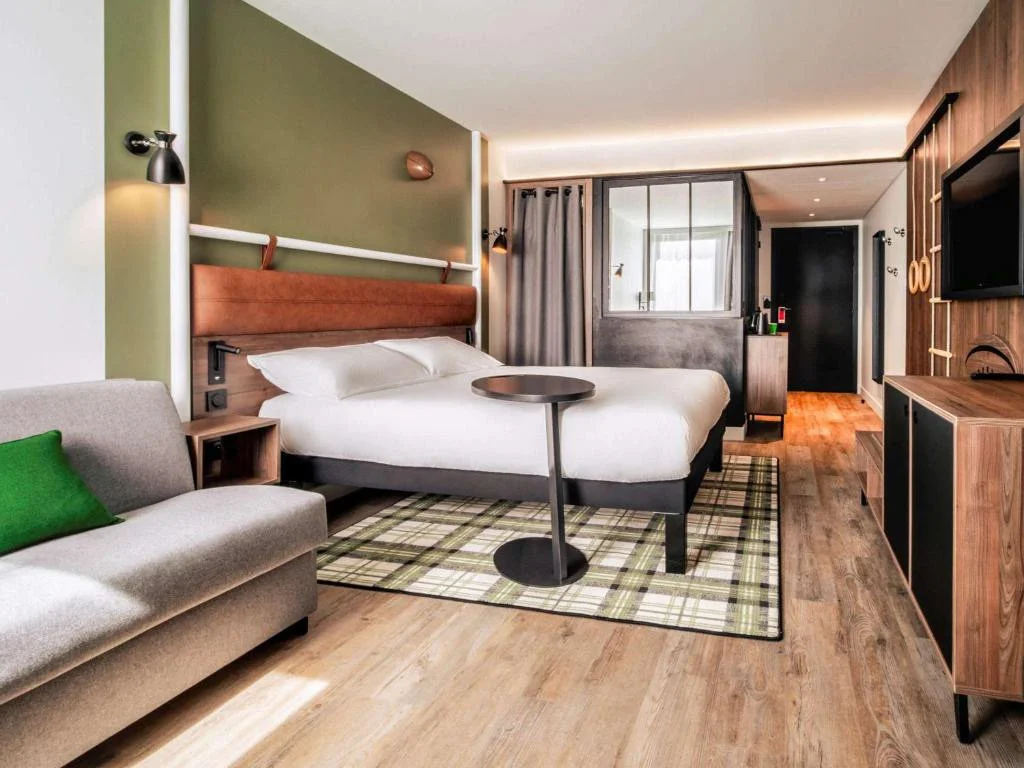 Une chambre d'hôtel moderne avec un lit, un canapé gris avec un coussin vert, un tapis à carreaux, un meuble TV en bois, une télévision, et une fenêtre avec des rideaux gris.