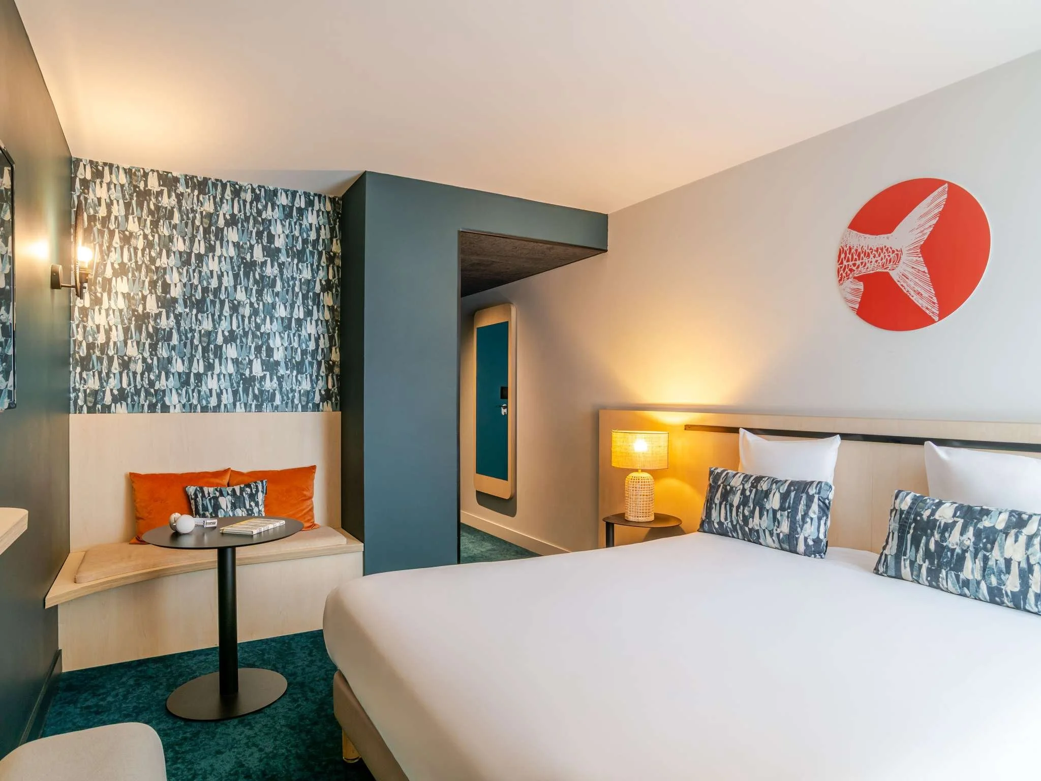 Une chambre d'hôtel moderne avec un lit blanc, des oreillers à motifs, une table ronde avec une lampe, une assiette avec des objets et un fauteuil orange. La décoration comprend un mur à motifs de plumes, un cercle rouge avec une étoile blanche en fo