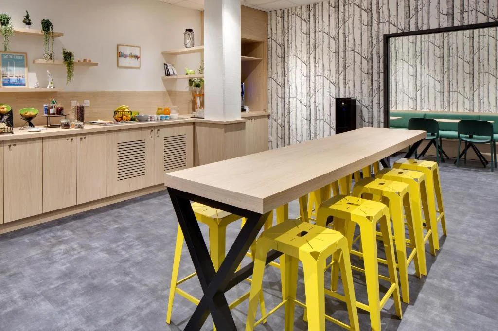 Salle avec comptoir de buffet, étagères avec plantes, murs en bois et papier peint arbre, table haute avec chaises jaunes, coin avec table et chaises vertes.
