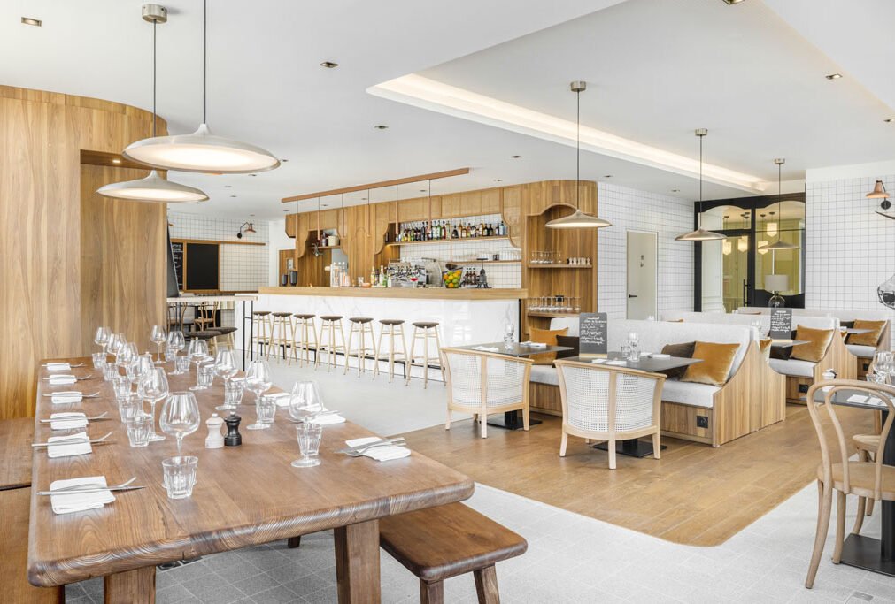 Intérieur d'un restaurant moderne avec un bar en bois, tables en bois avec assiettes et verres, et un coin salon avec des canapés et des chaises en bois.