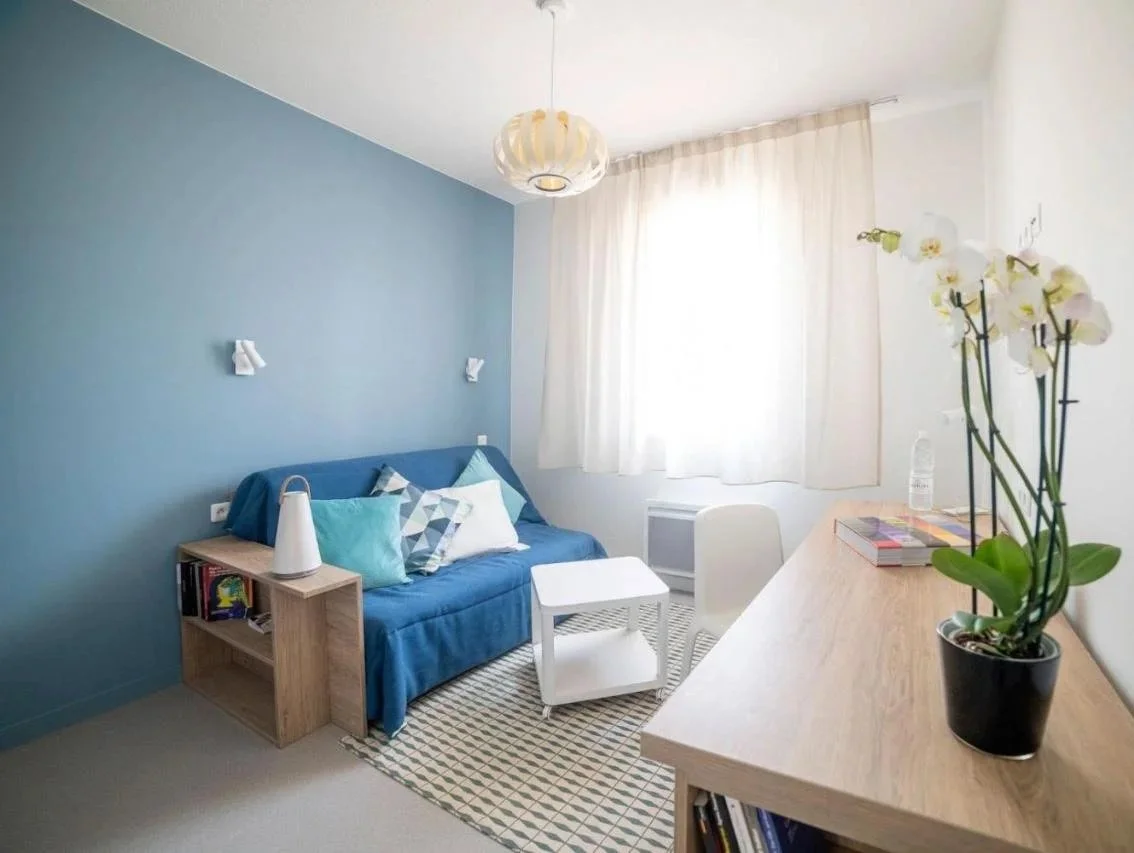 Salon avec un canapé bleu, des coussins, une table basse blanche, un lampadaire blanc, un mur bleu, et une étagère en bois avec des livres et une orchidée