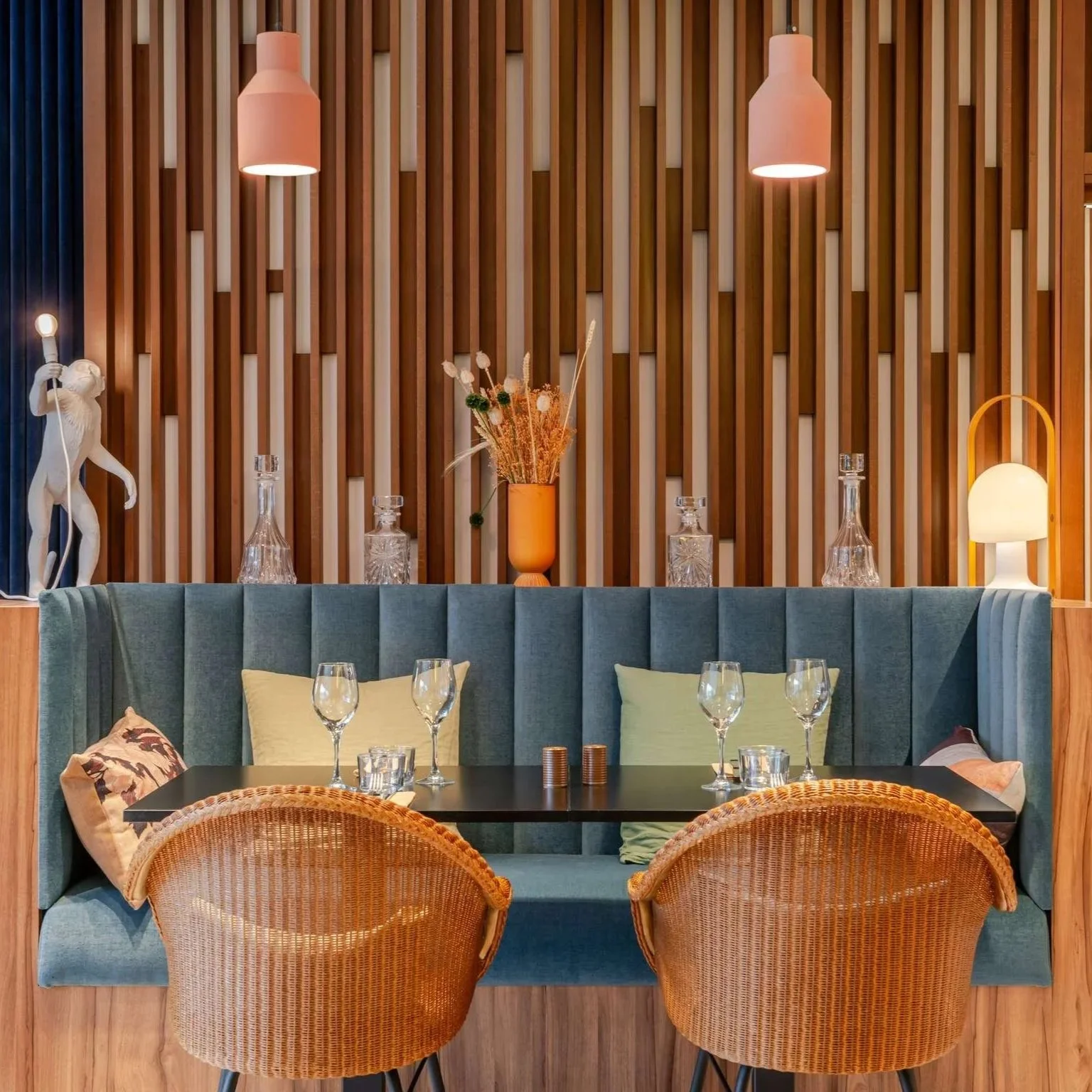 Une salle à manger élégante avec un mur en bois, une banquette rembourrée bleue, des fauteuils en rotin, une table avec des verres à vin, des coussins et des décorations florales sur une étagère, éclairée par des lampes suspendues et une lampe sur le côté.