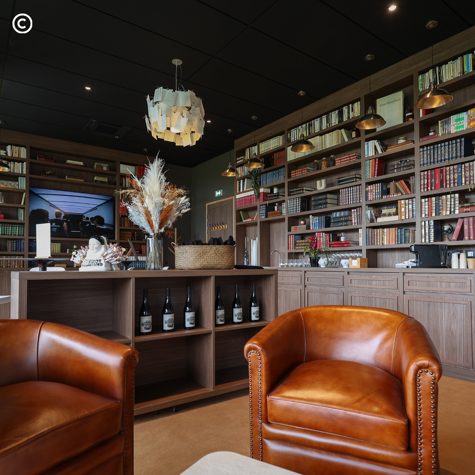 Intérieur d'une bibliothèque ou salon de lecture avec des étagères en bois remplies de livres, un grand écran, un centre de table avec des décorations et quatre fauteuils en cuir marron.