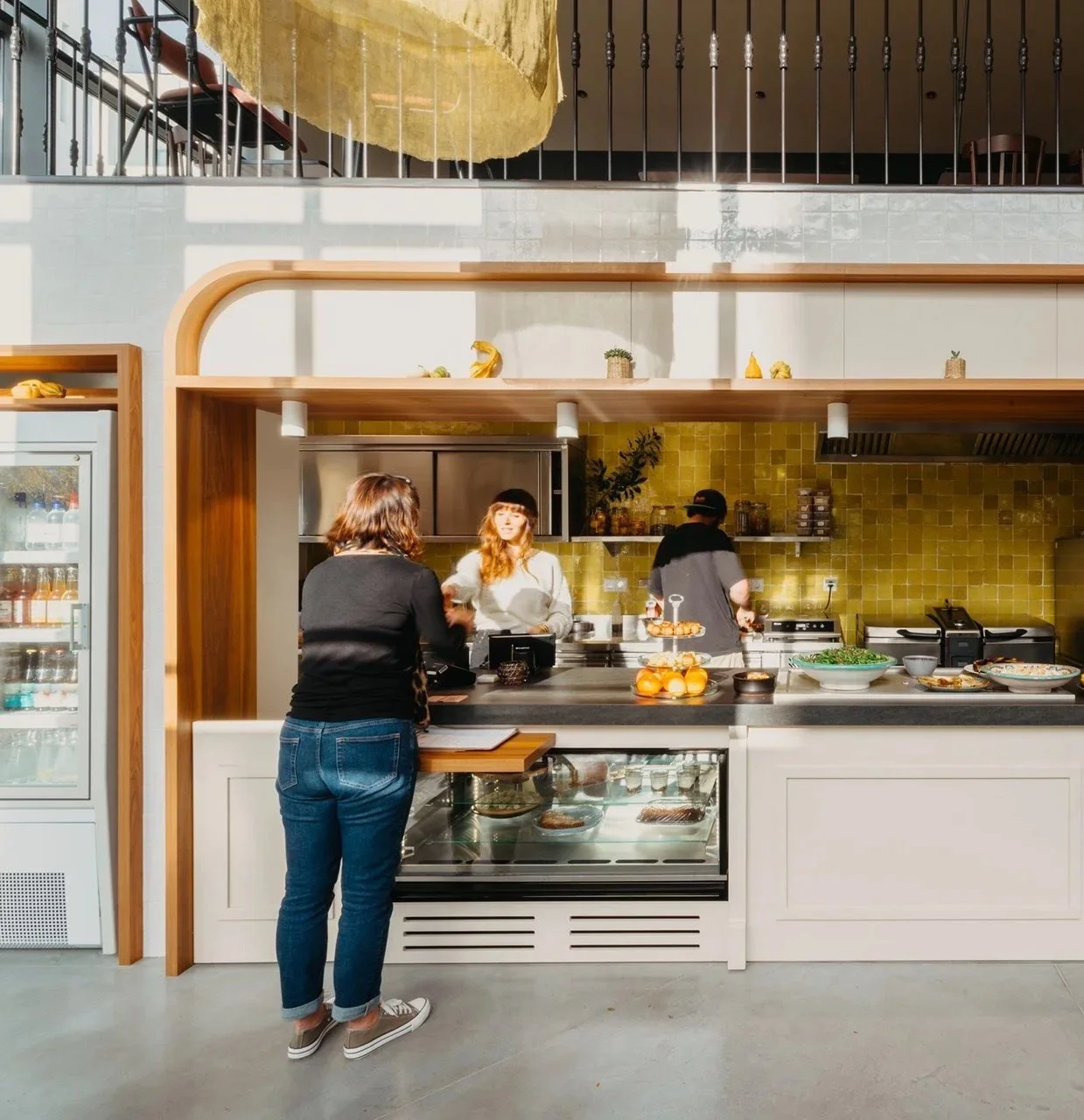Une femme parle avec deux employés dans une cuisine de restaurant, avec un comptoir en before, des fruits en décoration, et une étagère avec des plantes et des décorations.