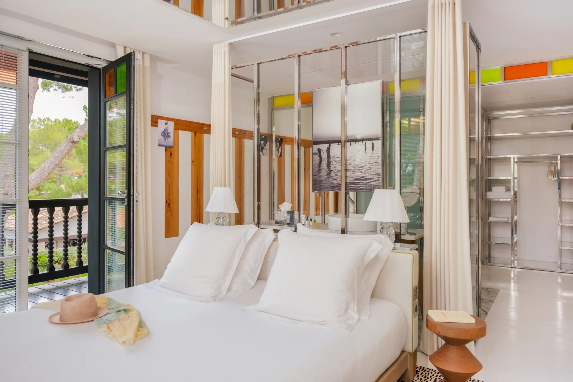 Chambre avec lit blanc, deux lampes de chevet, un miroir, une porte-fenêtre menant à un balcon, et des décorations en bois et verre avec des photos d'enfants jouant dans l'eau.