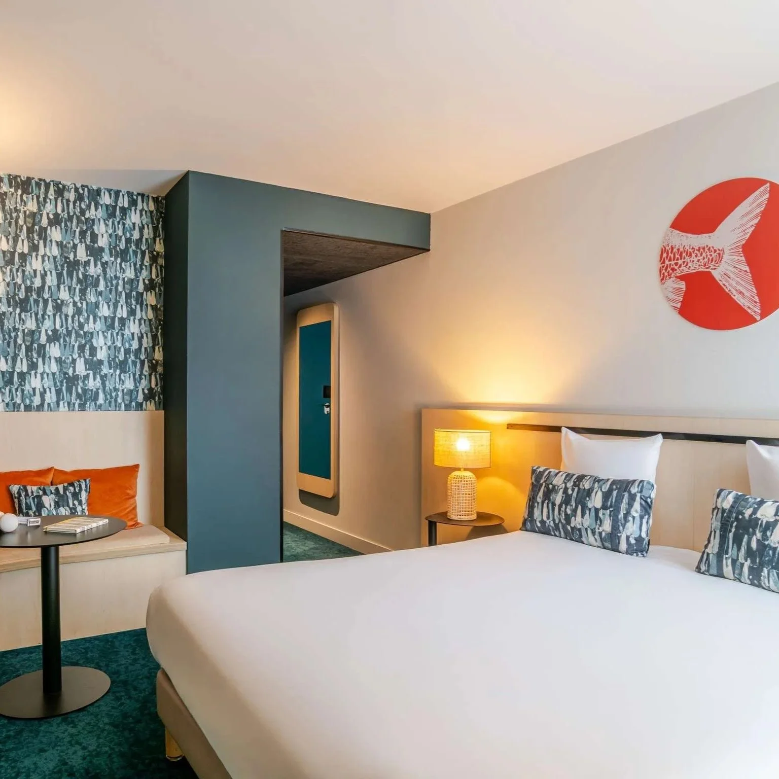 Chambre d'hôtel avec lit double, lampadaire, coussins avec motif d'oiseaux, décoration murale en forme de poisson, coin lecture avec fauteuil et table.