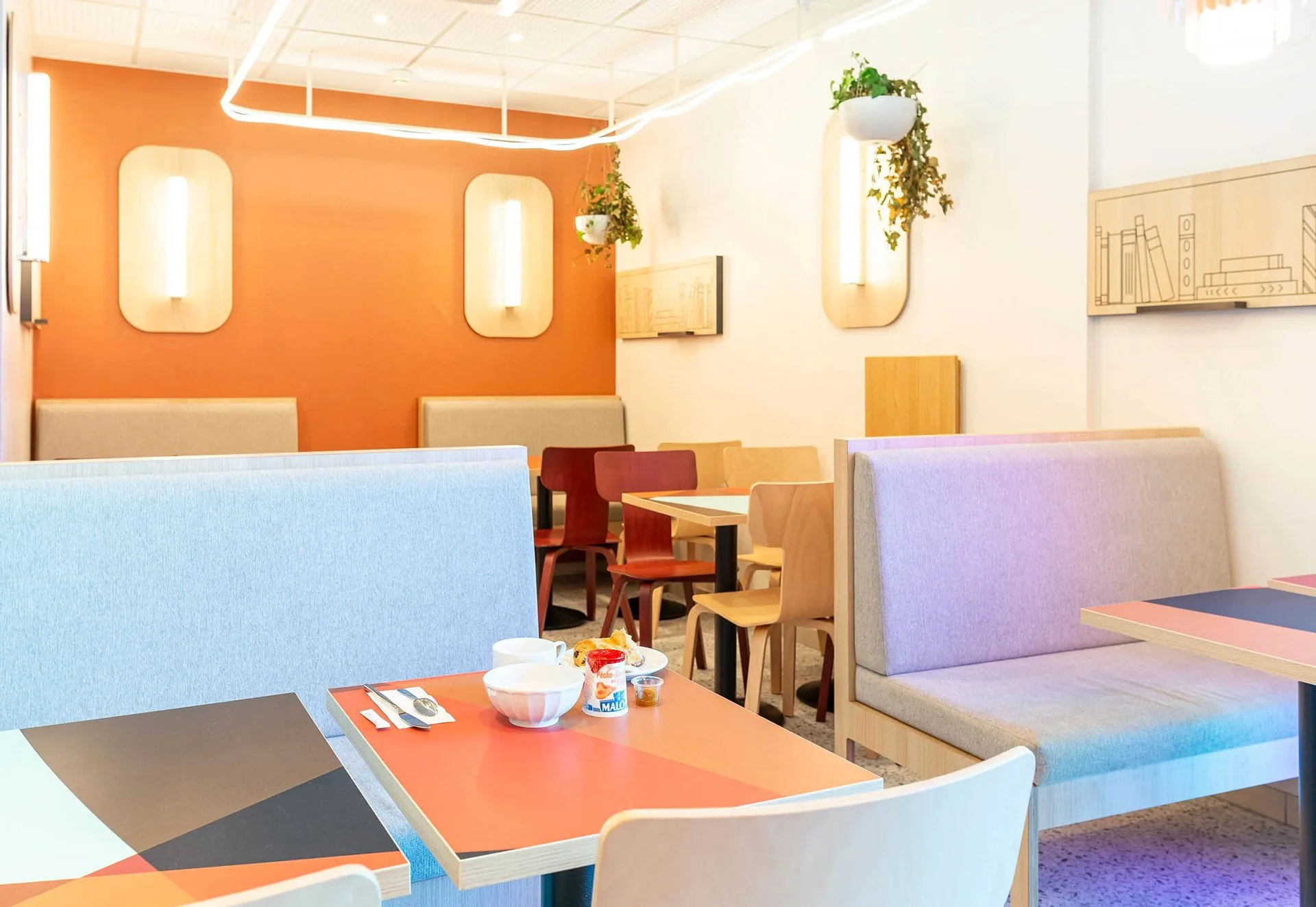 Intérieur d'un restaurant avec des tables, des chaises en bois, des banquettes, des plantes suspendues, des murs décorés, et un petit déjeuner avec un bol, une confiture et des couverts.