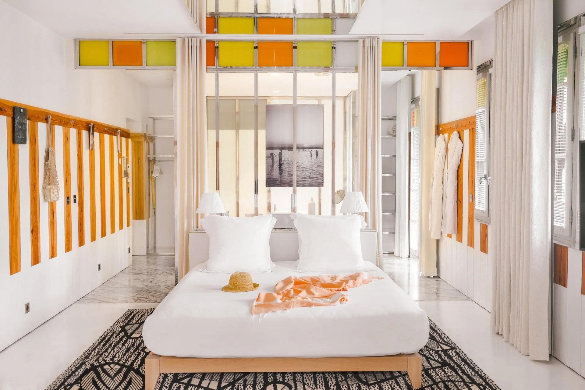 Chambre lumineuse avec lit king-size, décor blanc, touches de bois, fenêtres avec volets, tableau de plage au mur.