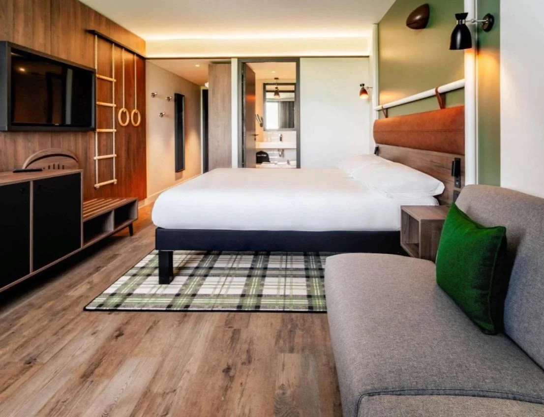 Chambre d'hôtel avec un lit double, un canapé gris avec un coussin vert, une télévision, et un mur en bois avec des équipements de sport, un tapis à carreaux au sol, et une salle de bains visible en arrière-plan.