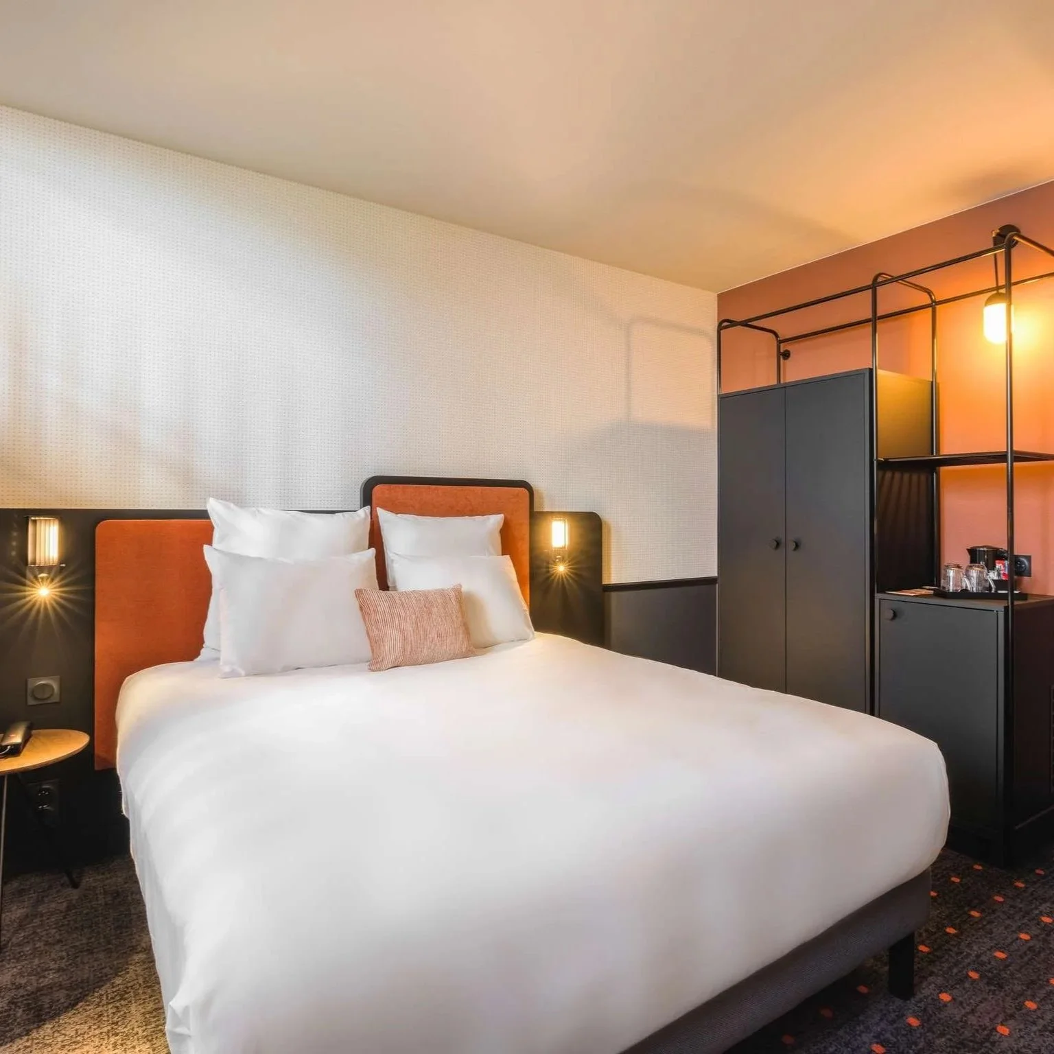 Chambre d'hôtel moderne avec un lit double, tête de lit orange, lampes de lecture, armoire noire et espace de service avec machine à café, ambiance chaleureuse.