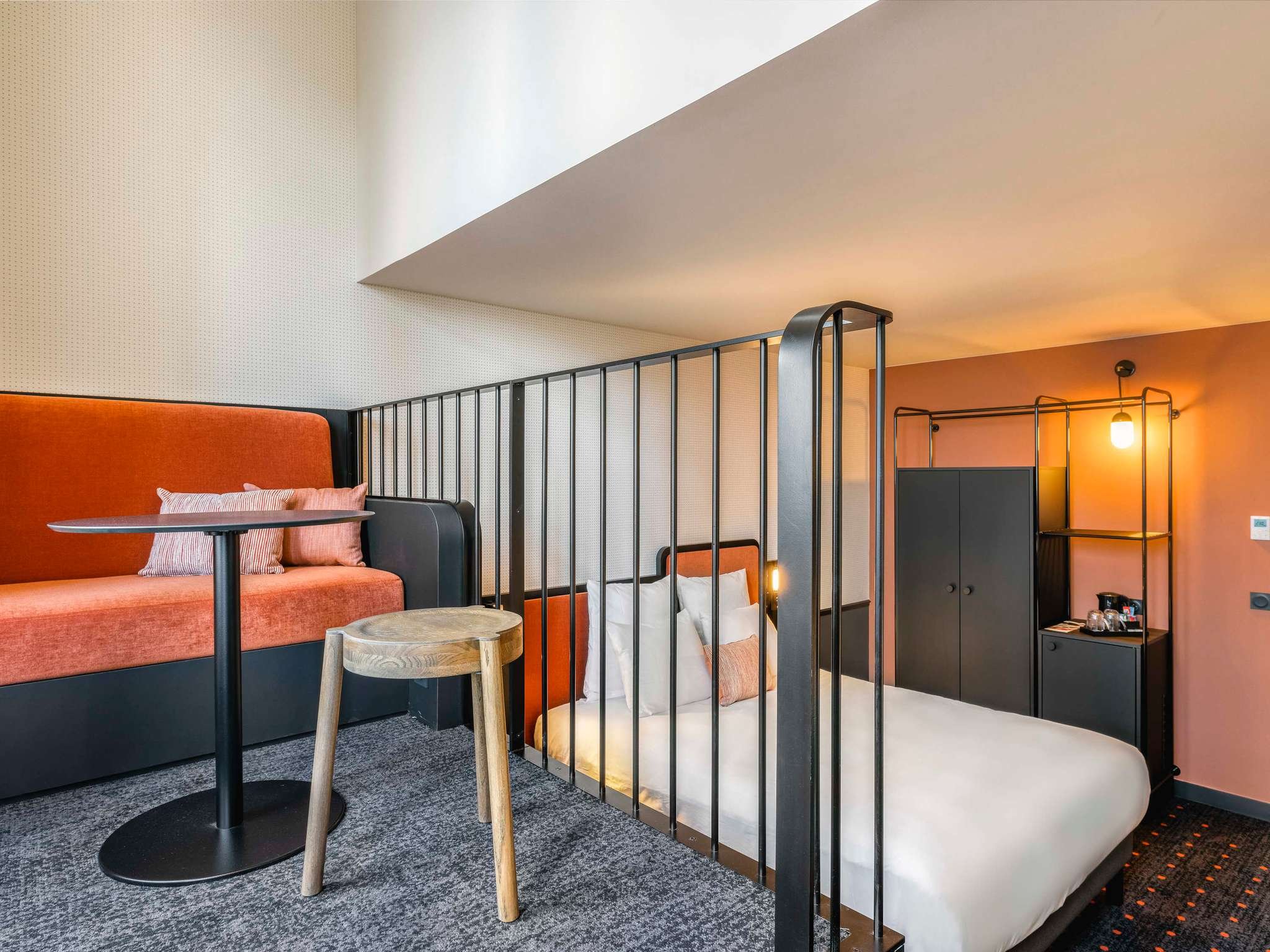 Intérieur d'une chambre ou d'un espace de vie avec un lit simple, une cloison en métal noir, un canapé orange avec coussins, une table ronde en bois, et une lumière chaude dans une pièce avec un mur orange.