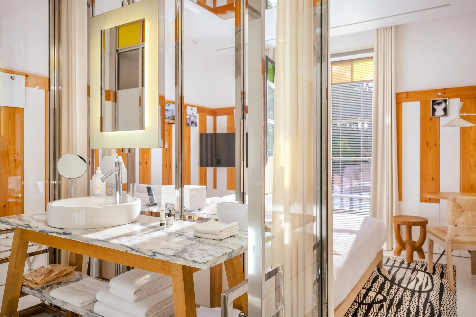 Salle de bain avec lavabo, miroir avec éclairage, et meubles en bois, vue à travers une cloison en verre avec rideau blanc, dans une pièce lumineuse avec fenêtres à volets.