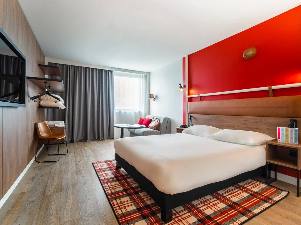 Chambre d'hôtel avec lit double, tapis à carreaux, paroi rouge, fenêtre avec rideaux gris, petit canapé avec coussins, television et espace de travail.