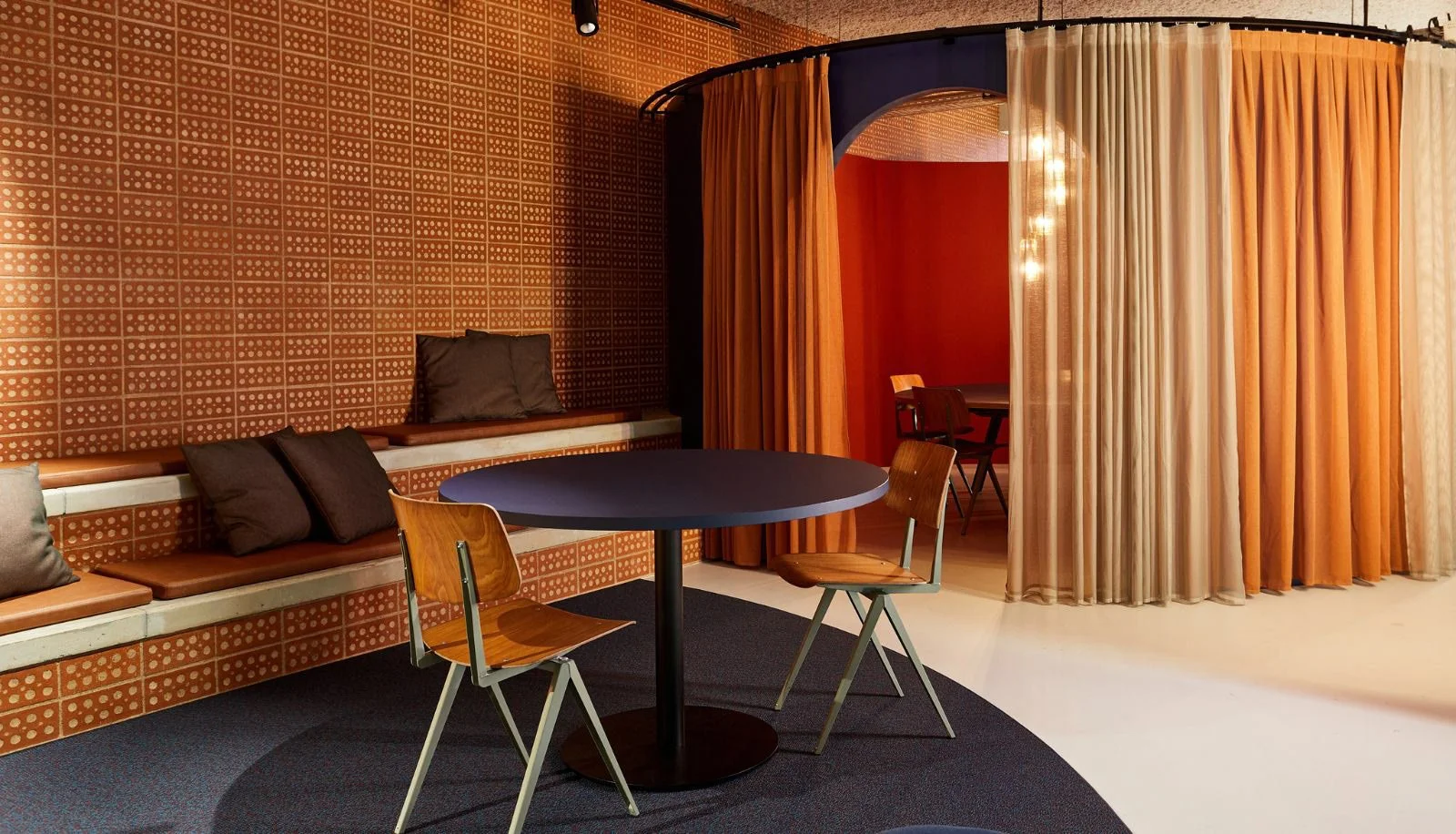 Intérieur d'une pièce avec un mur en brique orange, un canapé en cuir marron avec des coussins noirs, une table ronde noire avec deux chaises en bois, une cloison en rideaux orange et beige, une pièce derrière avec une table et des chaises.