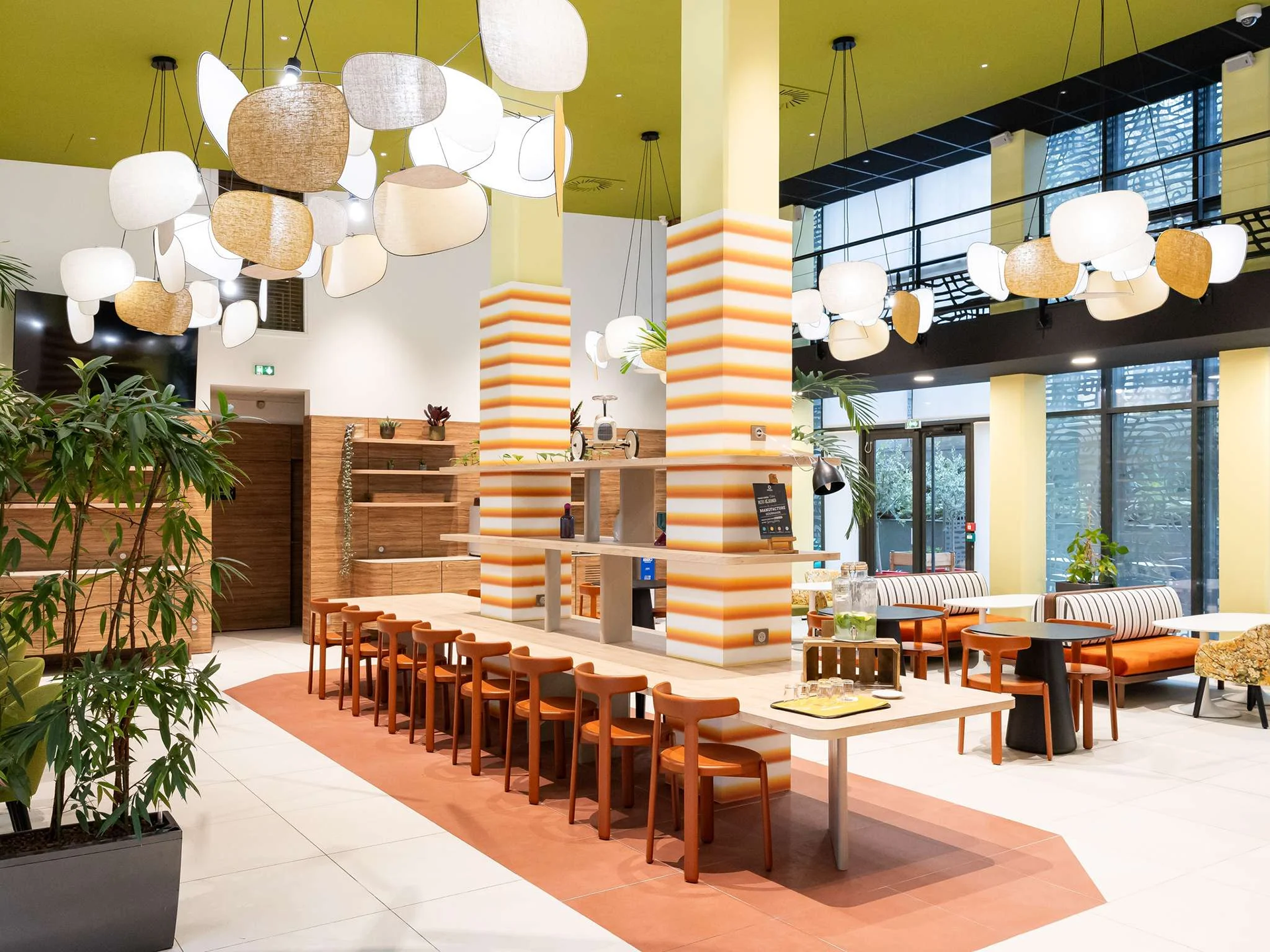 Salle de restaurant moderne avec un plafond vert, grands luminaires suspendus, murs blancs, mobilier en bois, plantes vertes, et grandes fenêtres laissant entrer la lumière naturelle.