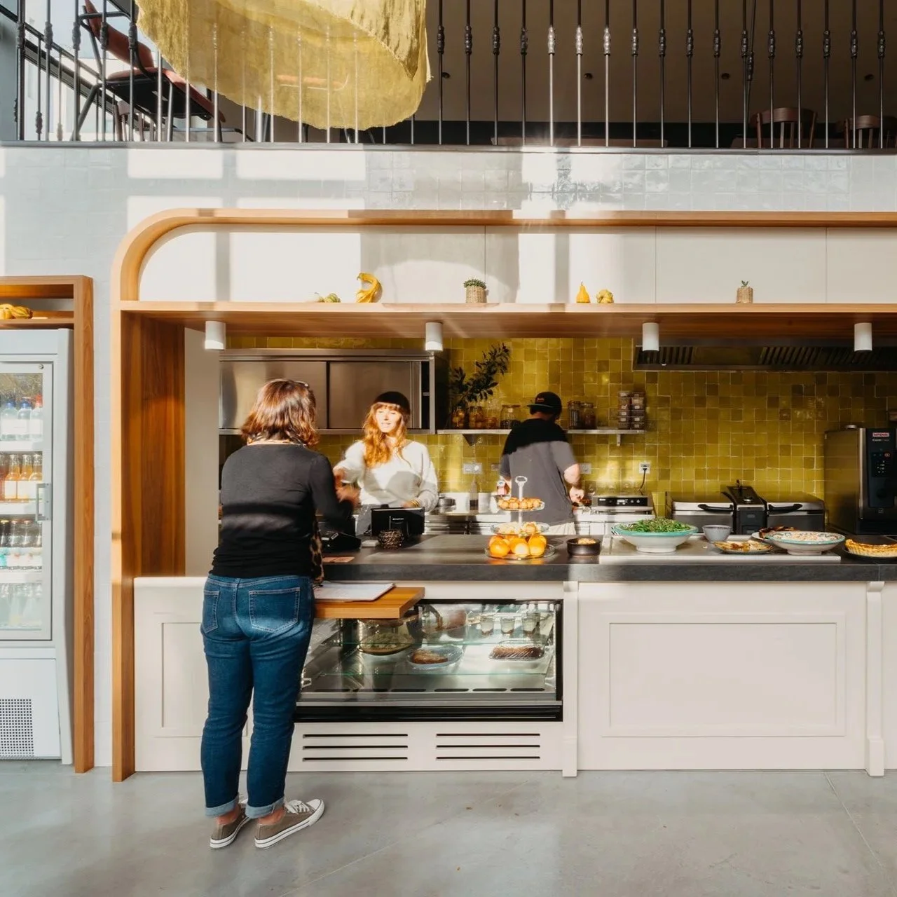 Une scène dans une cuisine moderne où deux femmes discutent avec deux cuisiniers. La cuisine a un mur en carrelage jaune, un comptoir noir, et des espaces de rangement en bois. Sur le comptoir, il y a des assiettes, des oranges, et diverses autres nourritures.