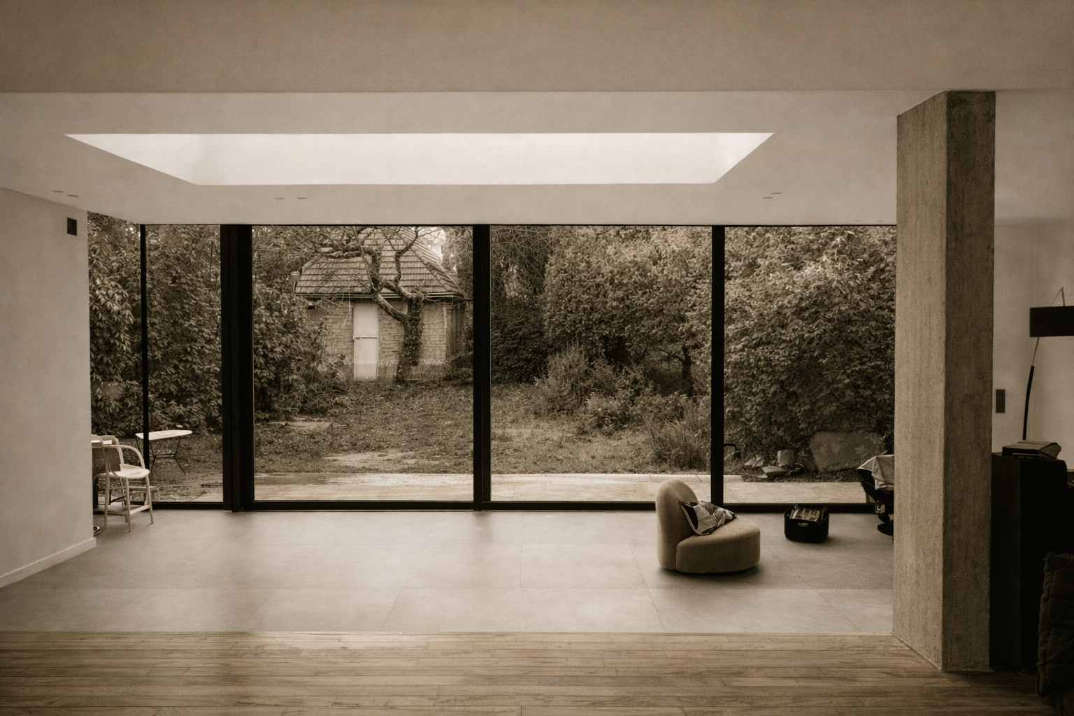 Intérieur d'une pièce moderne avec de grandes fenêtres vitrées donnant sur un jardin avec des arbres et un petit bâtiment. Meubles minimalistes, chaise ronde et une plante en fond.