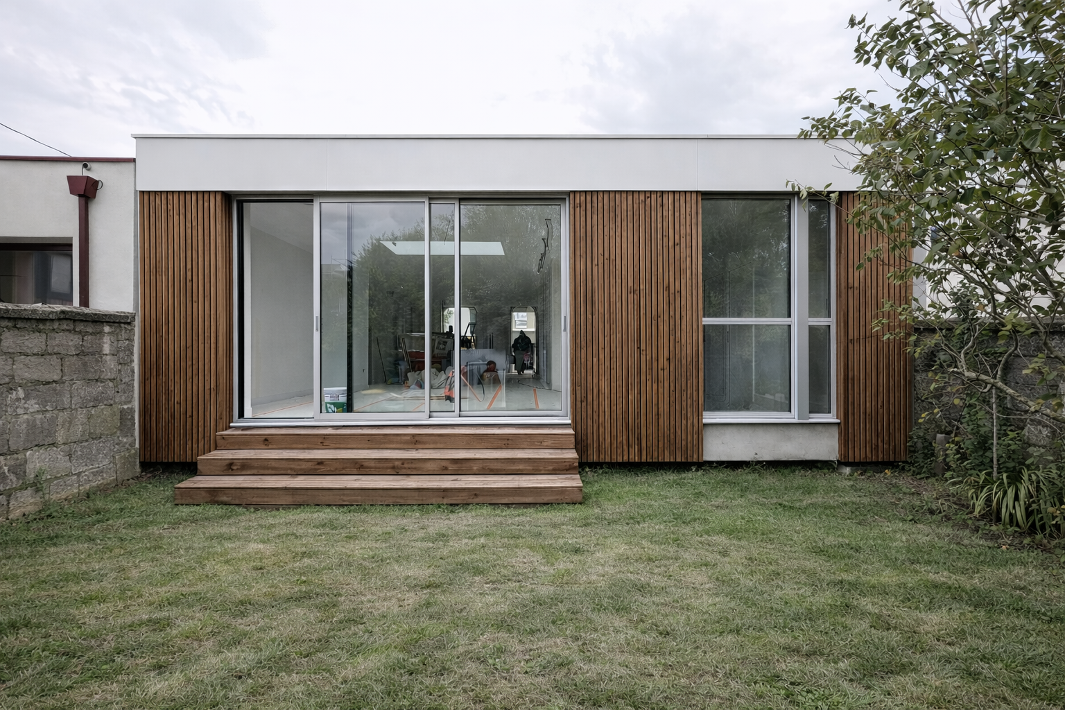 Une façade de maison moderne avec des murs en bois et de grandes fenêtres, y compris une porte-fenêtre en verre avec des marches en bois menant au jardin.