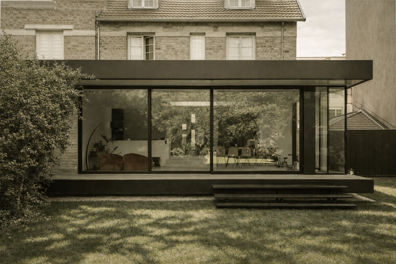 Une extension en verre moderne reliée à une maison traditionnelle à briques, avec des escaliers menant au jardin verdoyant