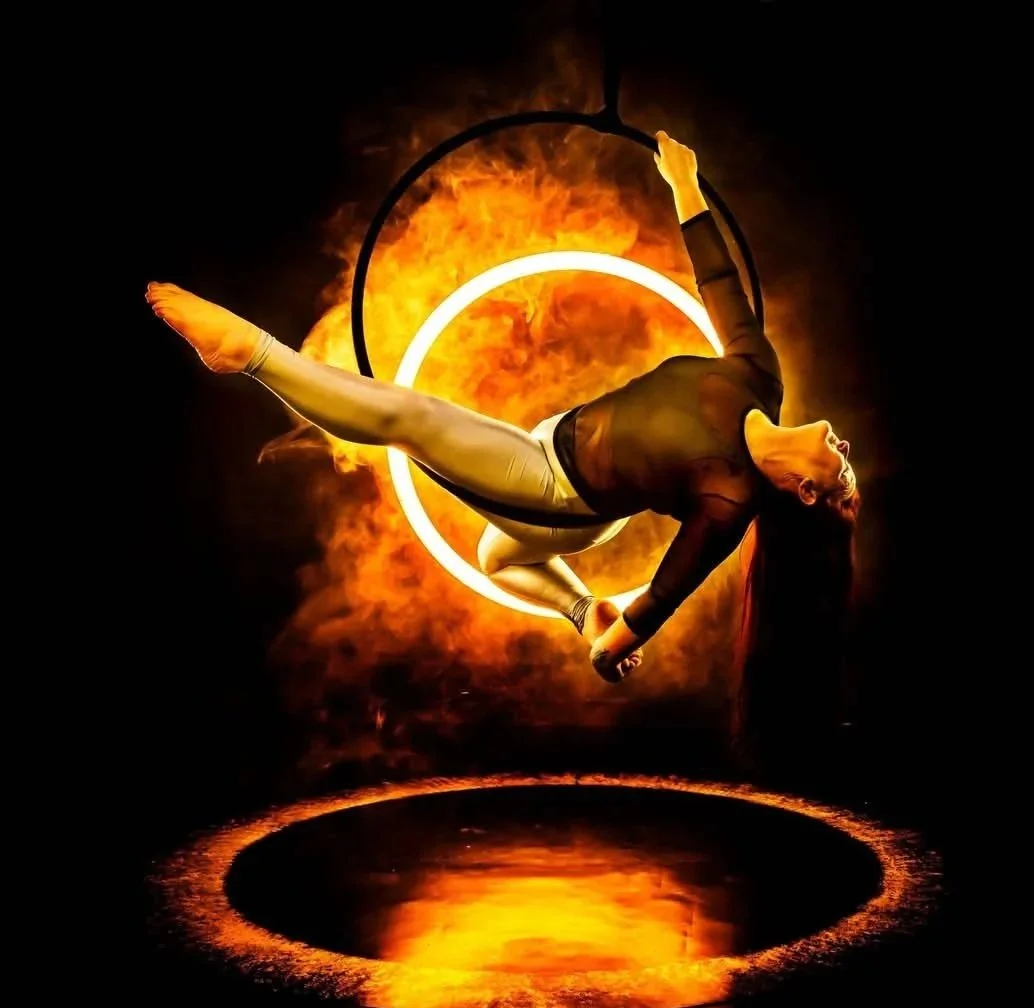 A female aerialist performing a pole act against a fiery background with glowing rings and smoke.
