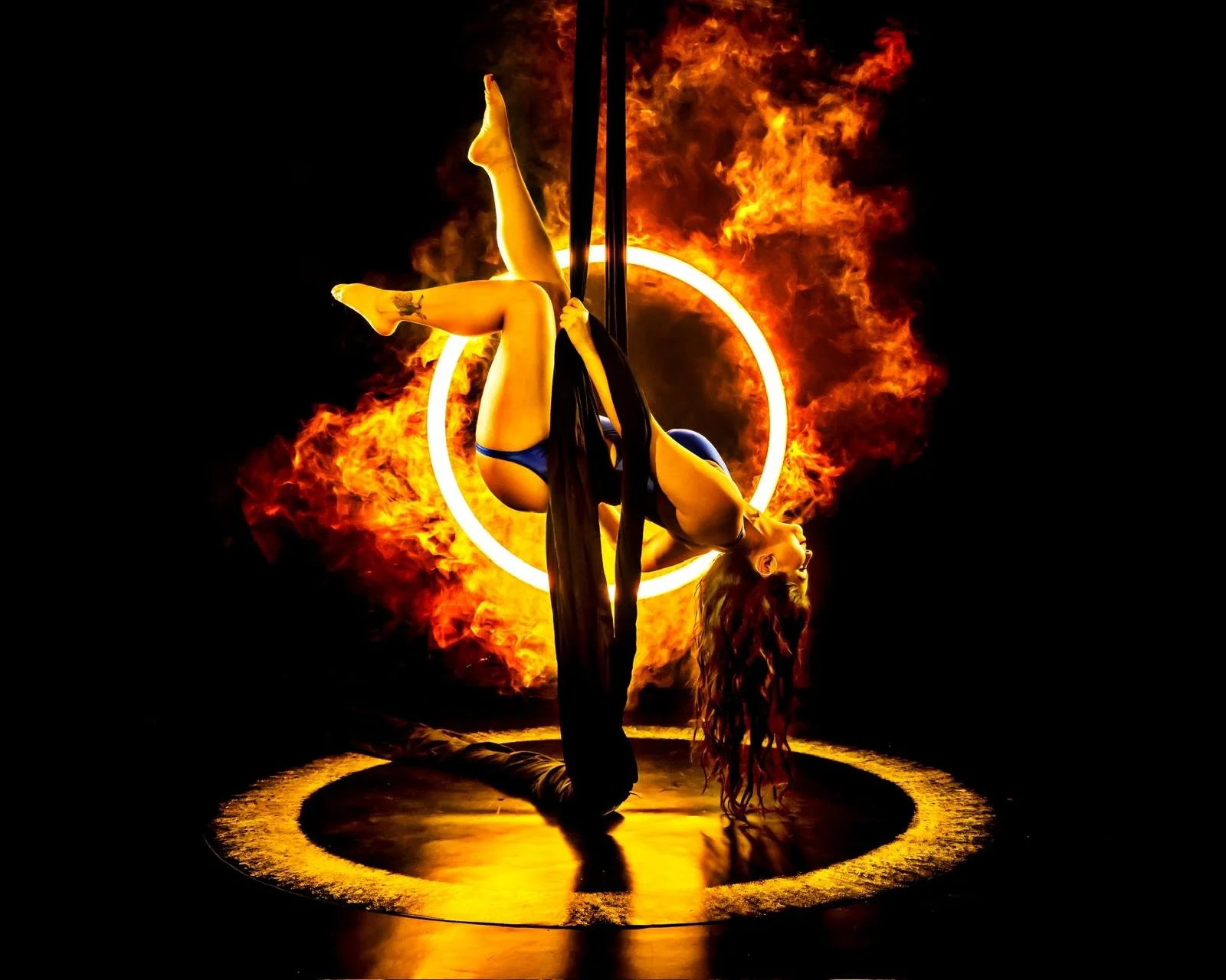 A female aerial performer hanging upside down on black silks against a backdrop of flames and a glowing circle, with a fire ring beneath her.