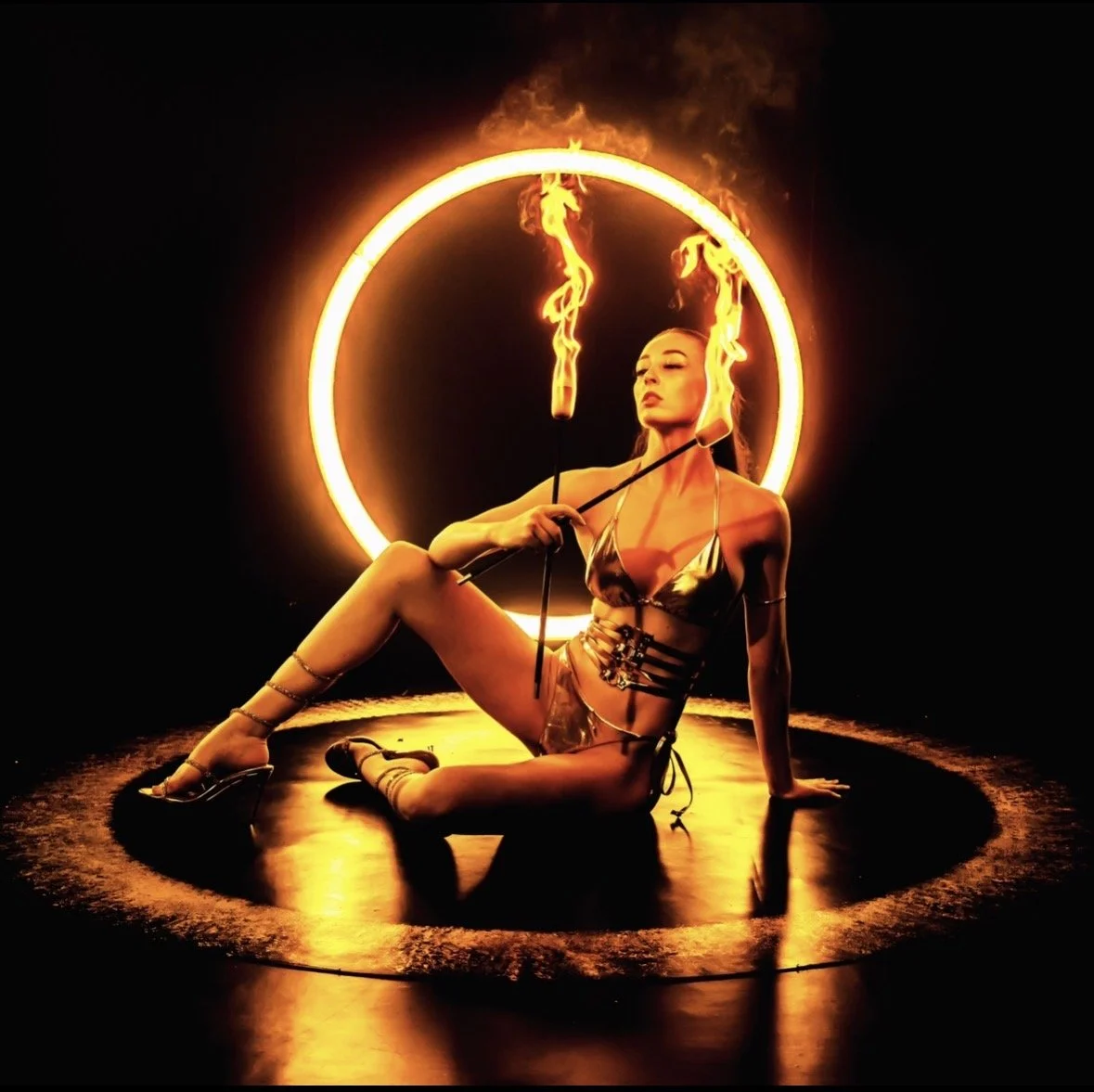 A woman sitting on the floor in front of a large illuminated circle with flames on a stick in each hand, posing amid flames and dark background.