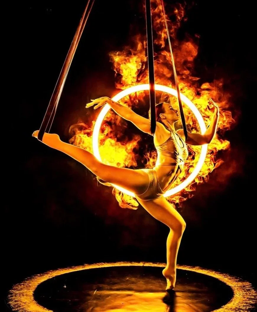 A female acrobat performing on aerial straps surrounded by flames and a glowing ring, with a dark background.