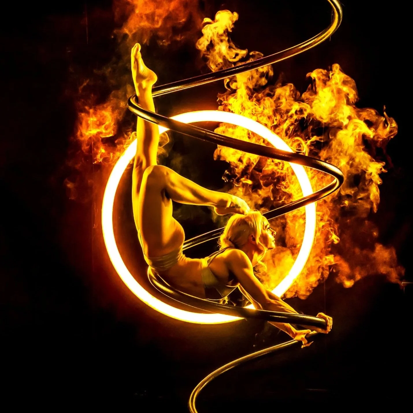 A female aerial performer caresses a flaming ring while surrounded by fire and smoke, performing a daring act against a dark background.